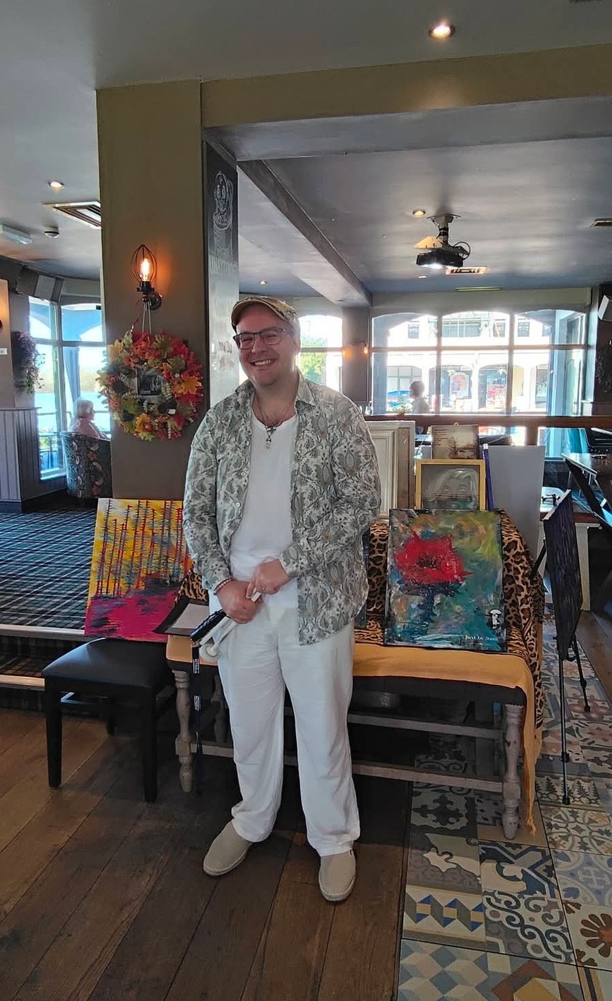Adrian Paternoster Adrian wearing white jeans, white tshirt, colourful shirt over the top in front of his artwork