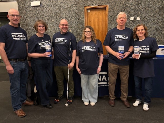 six volunteers standing in a semi circle wearing Retina UK blue tshirts with white RetinaUK emblems with collection boxes